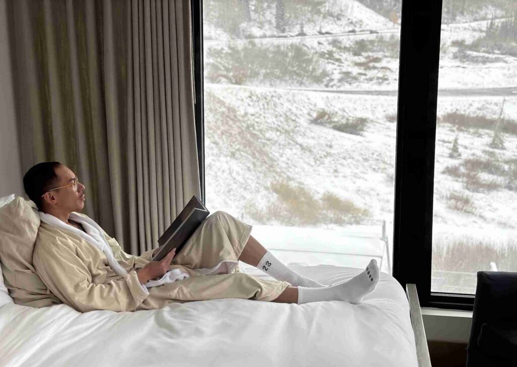 A person is relaxing in a steaming outdoor hot tub with a snowy mountain backdrop under a clear sky.