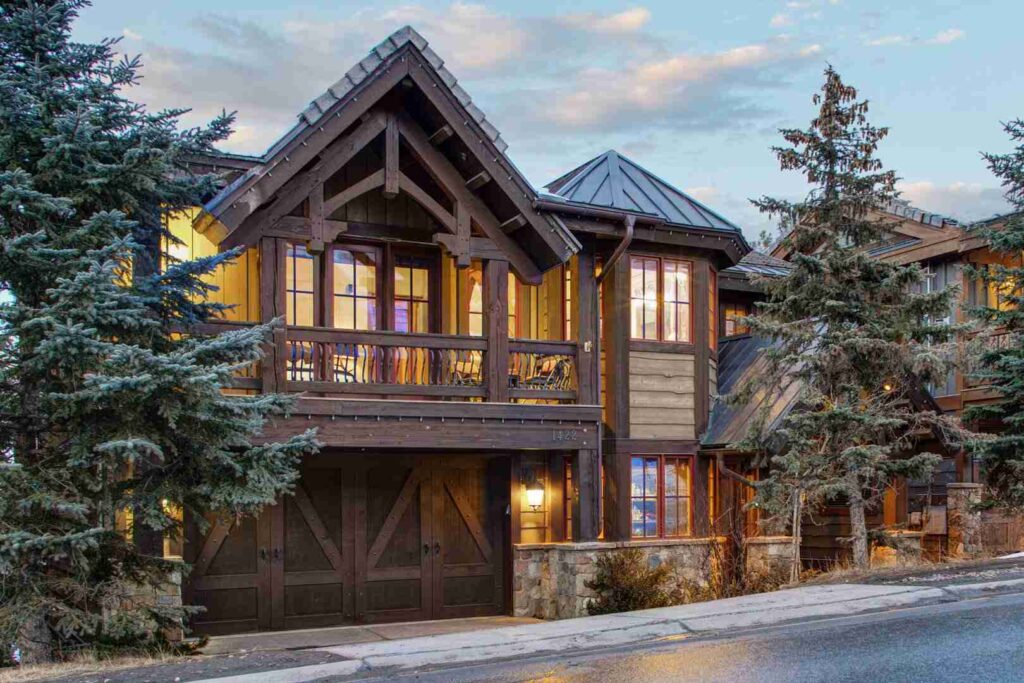 A large, illuminated wooden home with a stone foundation, surrounded by coniferous trees under a dusky sky.