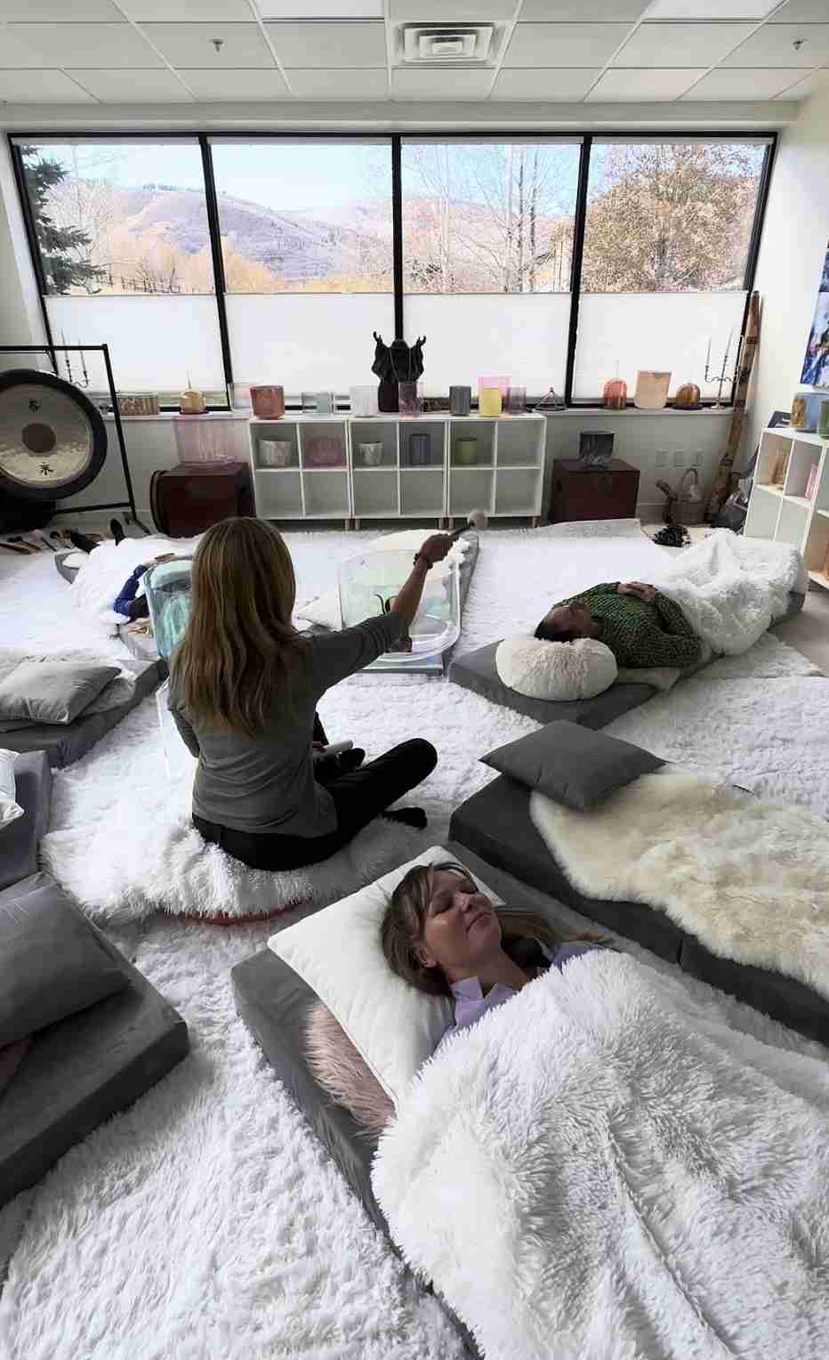 A group of people is lying on mats in a room with large windows offering a mountain view, participating in a relaxation or meditation session.
