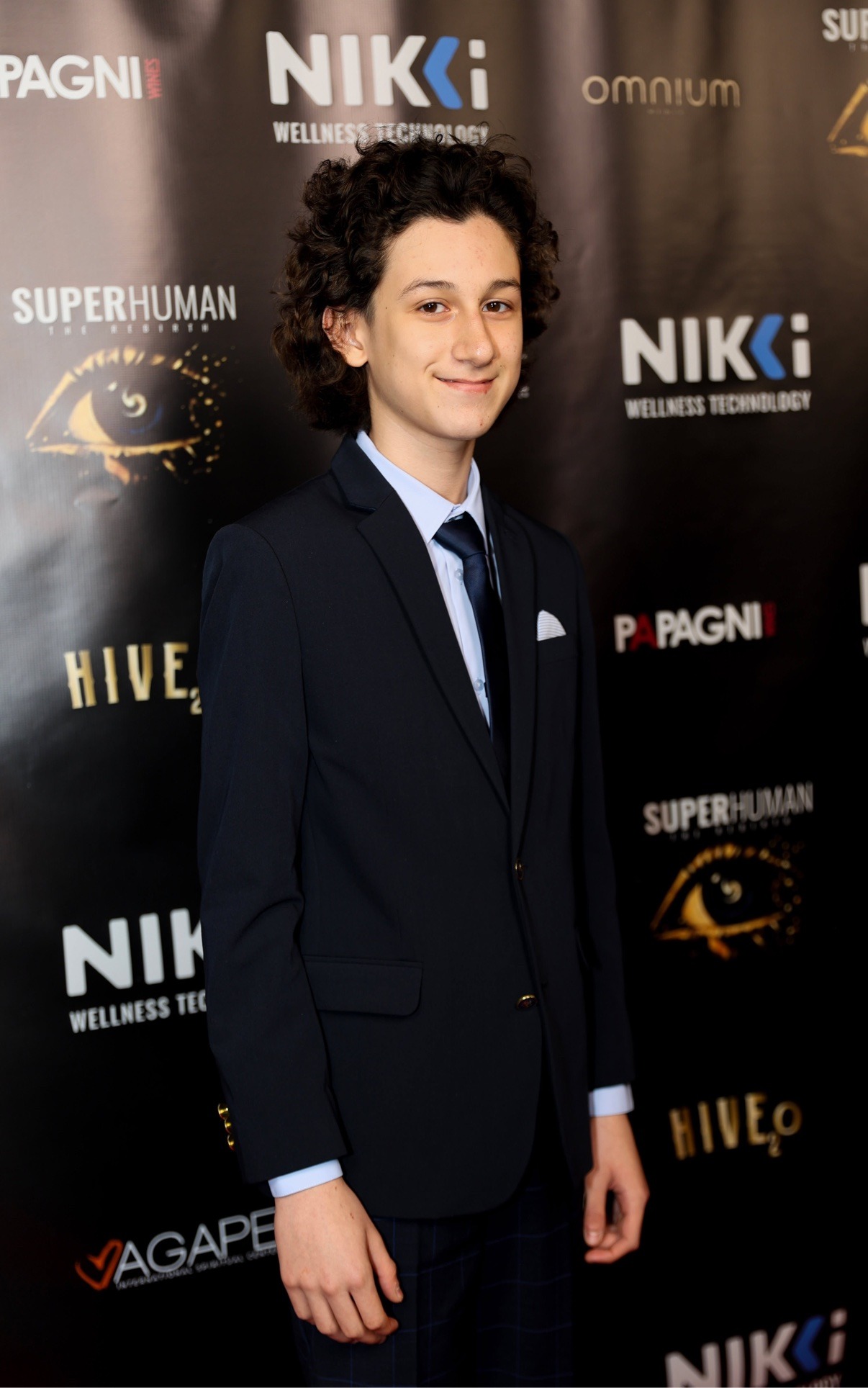 Young man with curly dark hair in a navy suit and tie on the red carpet at the Superhuman 2: The Rebirth premiere at the Saban Theatre, Beverly Hills