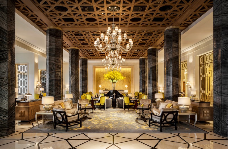 Grand lobby of a Four Seasons hotel in Dubai with a crystal chandelier, dark marble columns, patterned carpet and yellow accent chairs
