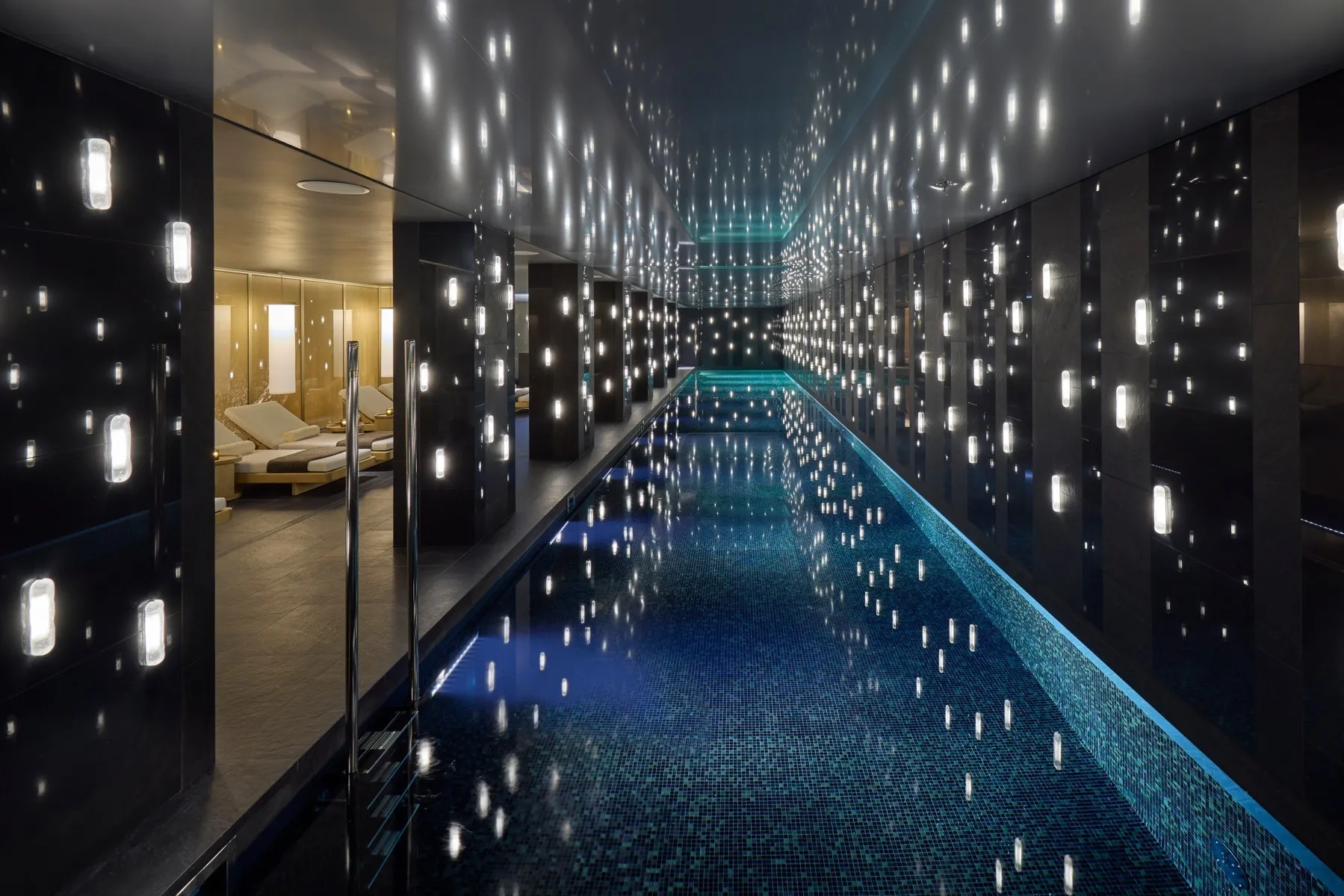 The indoor swimming pool at Mandarin Oriental Mayfair in London, a long dark tiled pool lined with glowing wall lights and lounge chairs