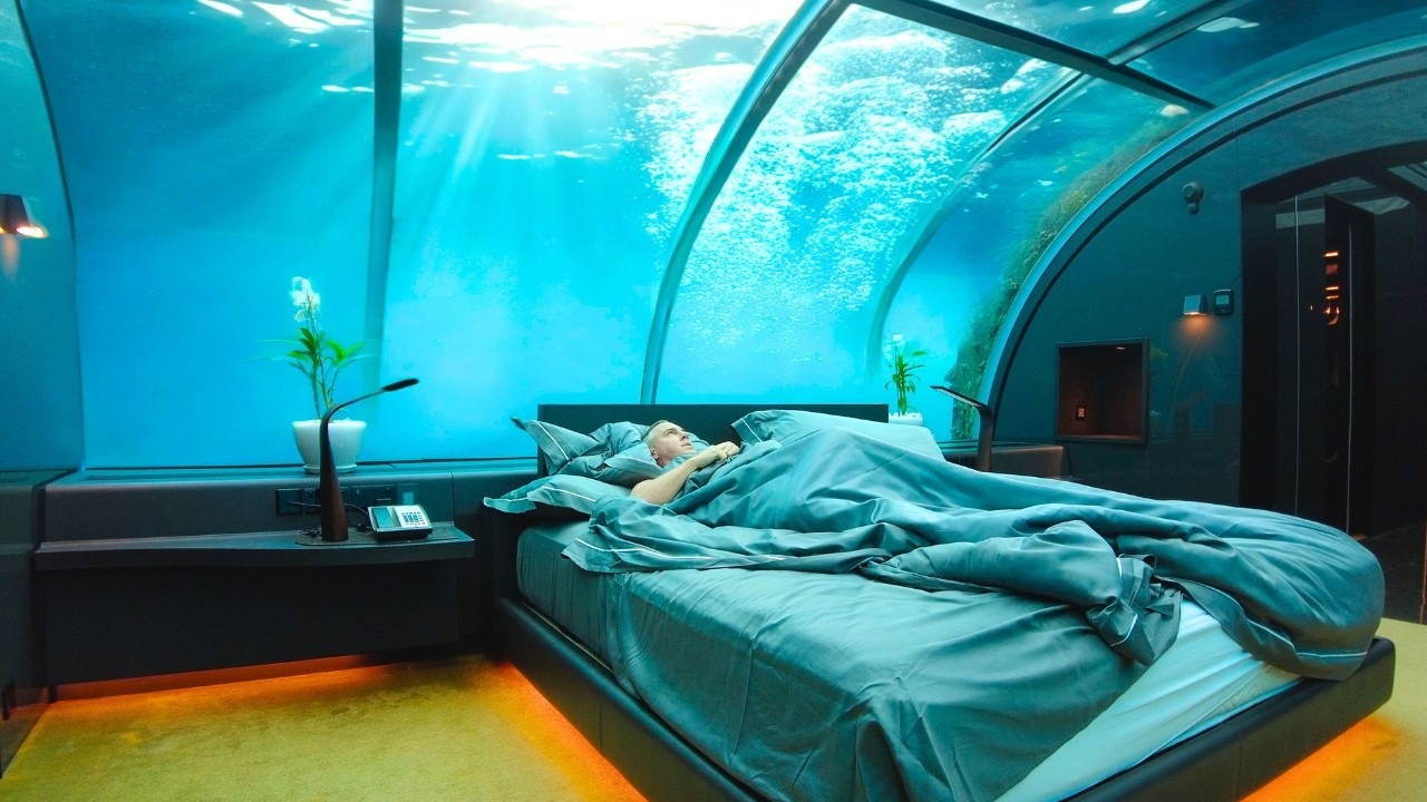 Walk With Me Tim lying in bed inside The Muraka underwater suite at Conrad Maldives Rangali Island, with the curved acrylic dome revealing blue ocean water overhead