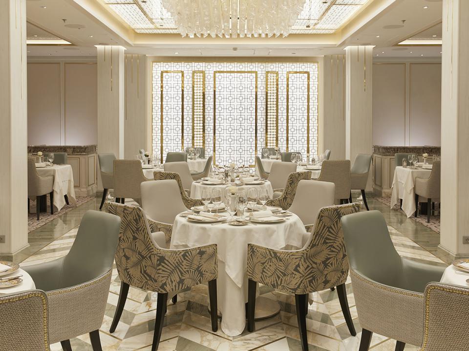 The Compass Rose main dining room aboard Seven Seas Splendor with set tables, upholstered chairs and gold patterned screens